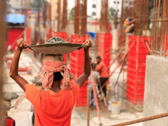 Photo of construction worker wearing orange t-shirt carrying building materials on a tray on her head.